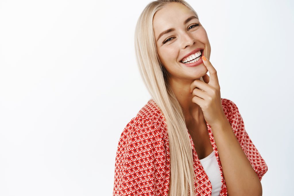 Beautiful blond girl with long healthy hair, laughing and smiling at camera, standing in summer clothes over white background