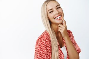 Beautiful blond girl with long healthy hair, laughing and smiling at camera, standing in summer clothes over white background